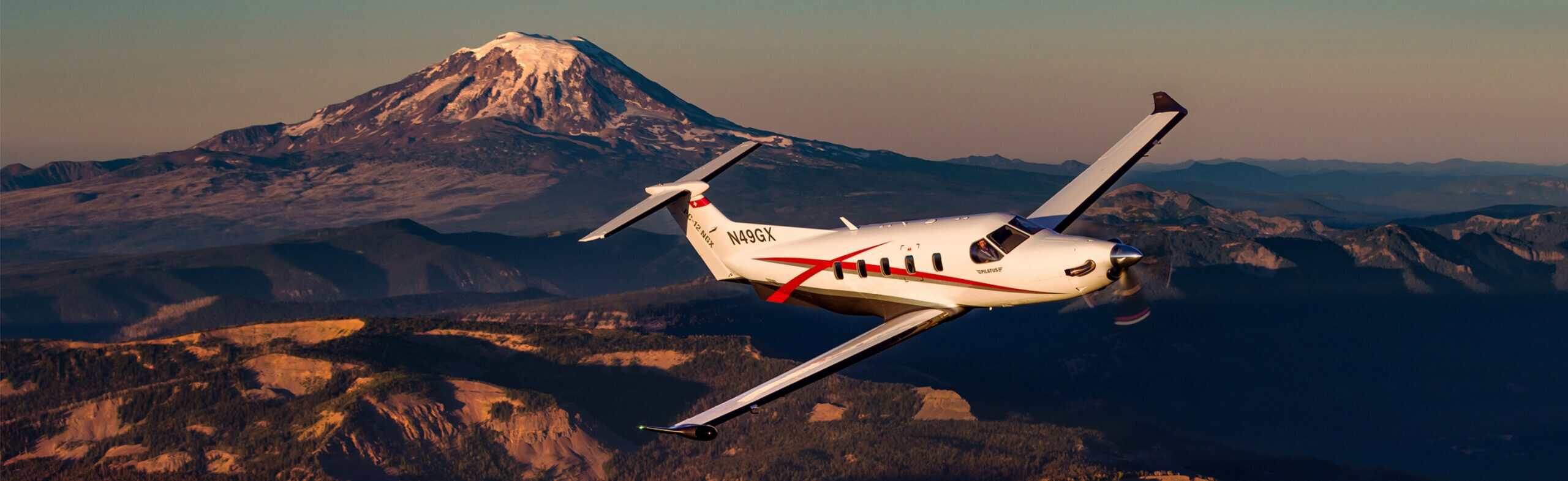 Pilatus PC-12 aircraft flying over mountainous terrain at sunset.